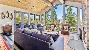 Un salon spacieux avec un grand canapé d’angle gris, une table basse et une cheminée. De grandes fenêtres offrent une vue sur les arbres et les montagnes.