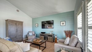 Ein Wohnzimmer mit blauer Wand, einem Holzschrank, einem beigefarbenen Sofa, einem Glastisch und einem Fernseher an der Wand.