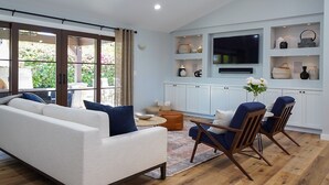 Uma sala de estar moderna com um sofá branco, almofadas azuis, uma mesa de centro de madeira e uma TV fixada na parede.