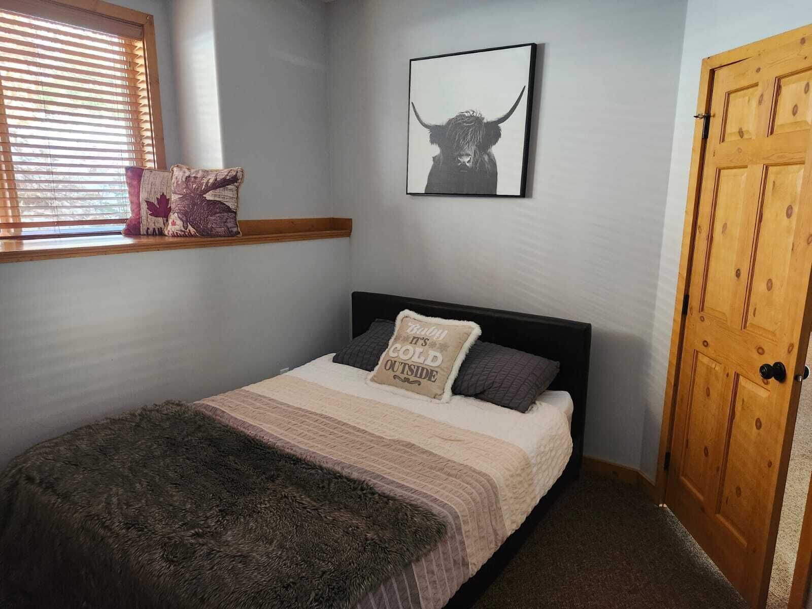 A bedroom with a bed, a window with blinds, a framed picture on the wall, and a wooden door.