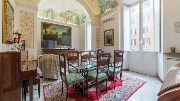 A dining room with a glass table, green upholstered chairs, a red patterned rug, and a large landscape painting on the wall.
