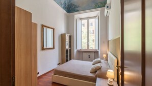 A bedroom with a bed, bedside table, lamp, window, and a ceiling mural.