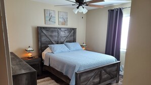 A bedroom with a wooden bed frame, a headboard, two wall-mounted lamps, a ceiling fan, and a window with curtains.