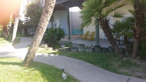 A pathway leading to a building with a covered porch, outdoor seating, and palm trees.