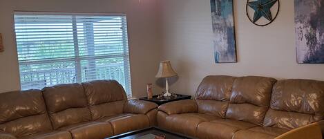 A living room with a brown leather sectional sofa, a glass-top coffee table, a ceiling fan, and artwork on the walls.