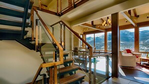 A modern interior with a glass staircase, wooden beams, and a view of mountains through large windows. - Stewart Highlands Vacation Home at Windcliff (Estes Park)