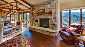 A spacious living room with a stone fireplace, wooden beams, and a large window offering a mountain view. - Stewart Highlands Vacation Home at Windcliff (Estes Park)