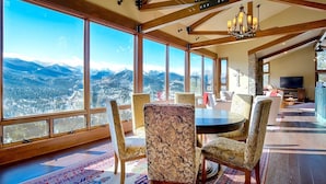 A spacious dining area with large windows offering a mountain view, wooden beams on the ceiling, and a chandelier. - Stewart Highlands Vacation Home at Windcliff (Estes Park)