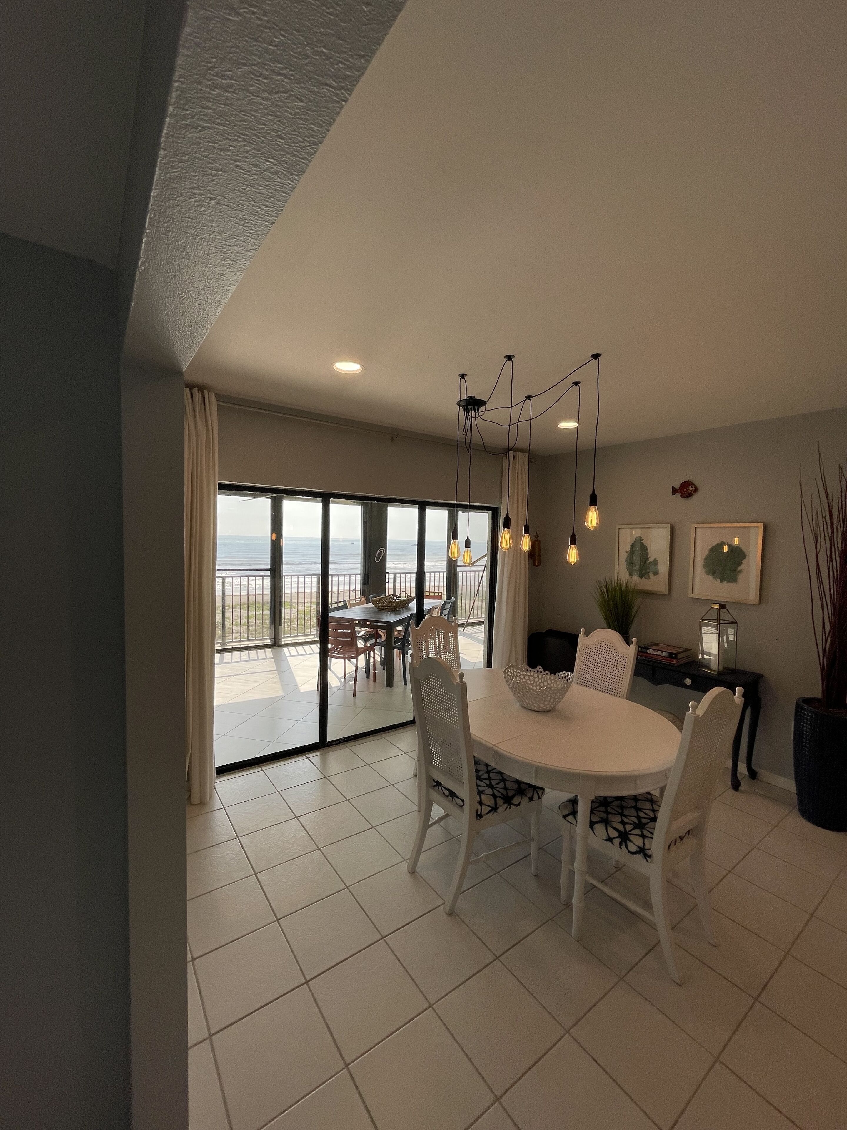 A dining area with a large table and chairs, a chandelier, and a view of the sea through large glass doors.