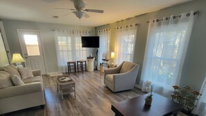 Un salon comprenant un canapé, un fauteuil, une table basse et un téléviseur. On y trouve également deux chaises, une lampe et une petite table sur laquelle est posé un objet décoratif.