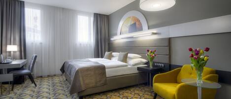 A hotel room with a bed, a desk, a chair, a yellow armchair, and a vase of flowers.