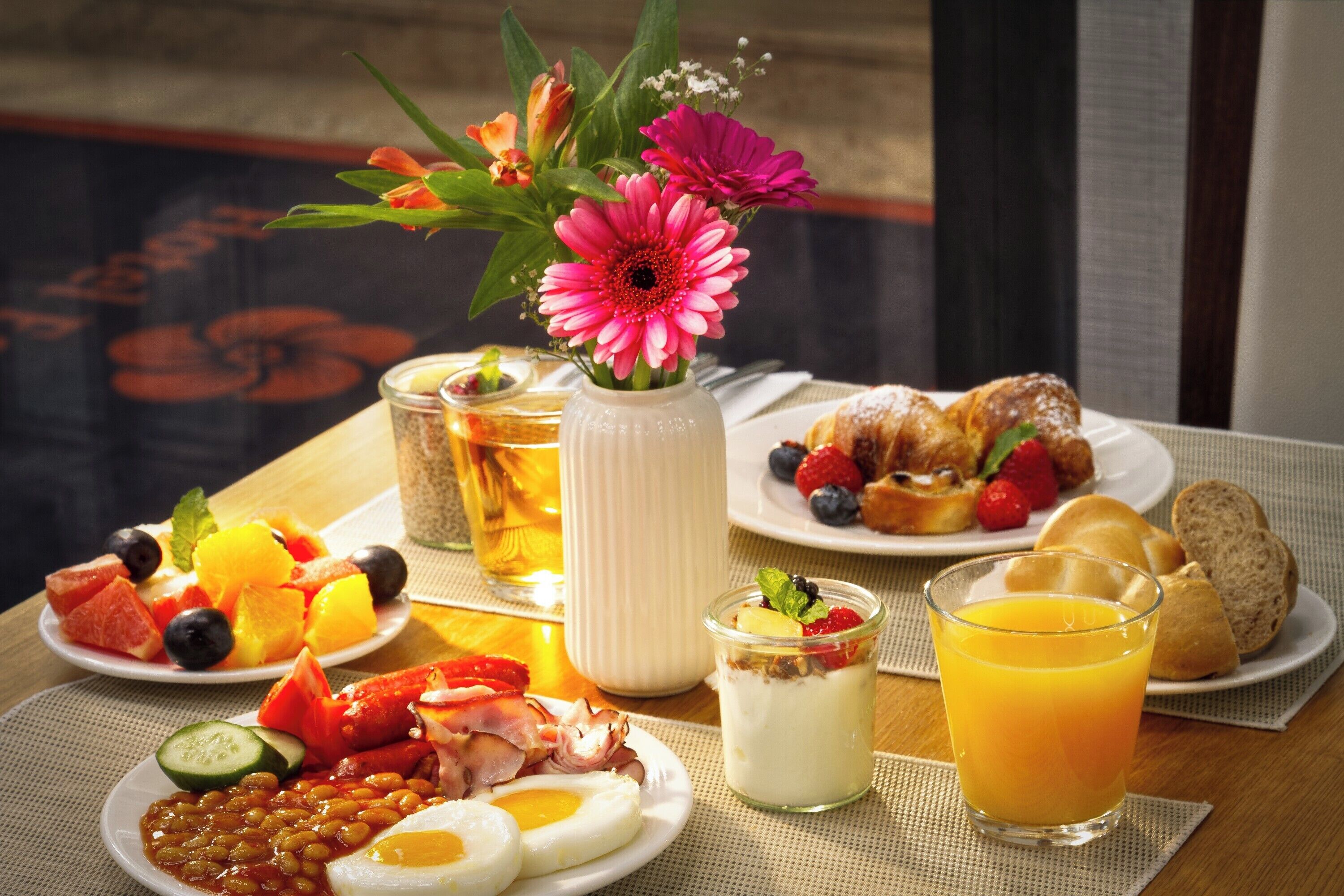 Una mesa de desayuno con diferentes platos, un vaso de jugo de naranja y un jarrón con flores.