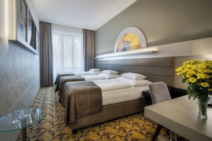 Quarto de hotel com chão estampado em padrão geométrico, cama com cabeceira, mesa de cabeceira com um vaso de flores amarelas e janela com cortinas.