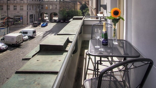 A balcony with a table and chairs, overlooking a street with a historic building and a clock tower in the background. - Hotel Essence  (Prague)