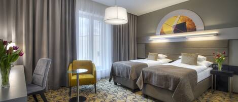 Habitación de hotel con dos camas, un escritorio, una silla y un amplio ventanal con cortinas.