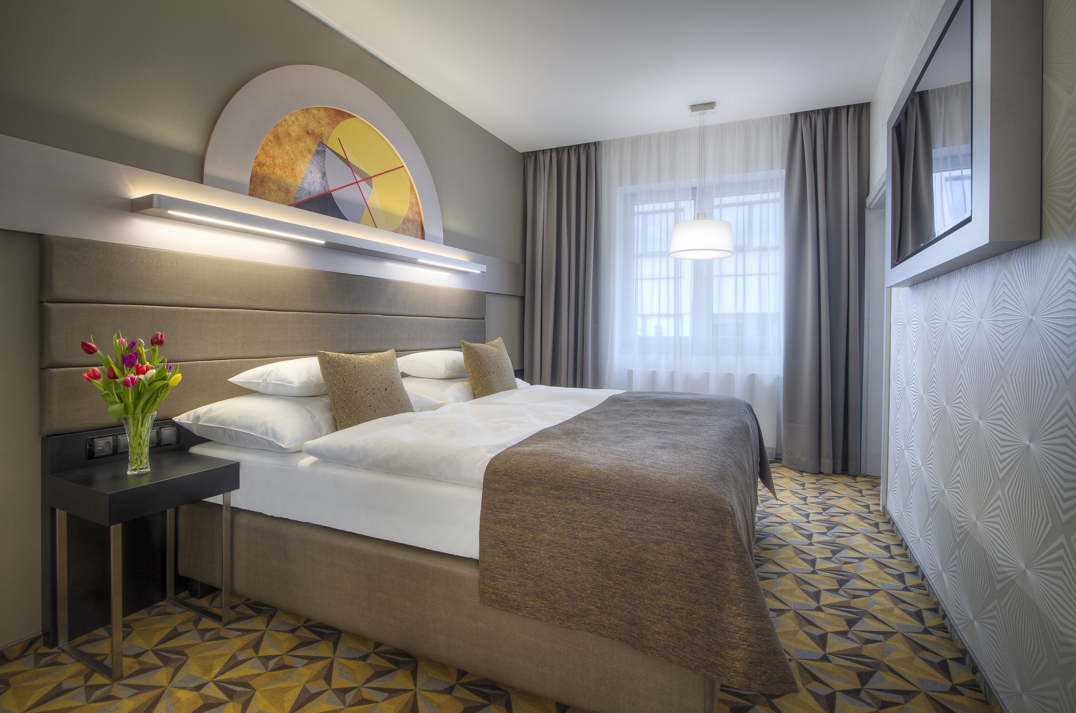 A hotel room with a large bed, a bedside table with a vase of flowers, and a window with curtains.