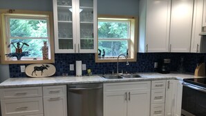 A modern kitchen with white cabinets, a stainless steel dishwasher, a double sink, and a large window with a view of trees. - Blue Mountain Cottage near Deep Creek Campground (Bryson City)