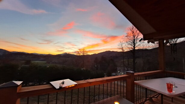 A deck with a fire pit and a table, overlooking a scenic landscape with hills and a sunset. - Blue Mountain Cottage near Deep Creek Campground (Bryson City)