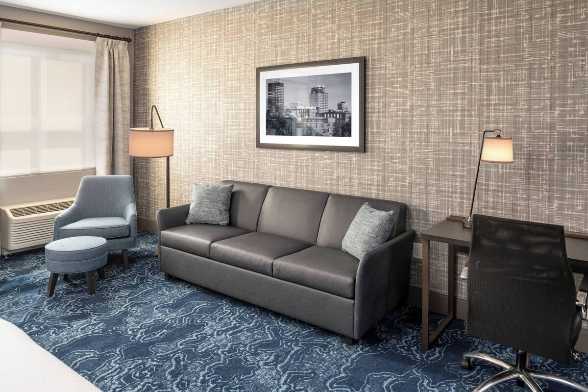 A hotel room with a grey sofa, a small round ottoman, a desk with a chair, and a framed cityscape picture on the wall.