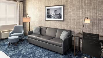 A hotel room with a grey sofa, a small round ottoman, a desk with a chair, and a framed cityscape picture on the wall.