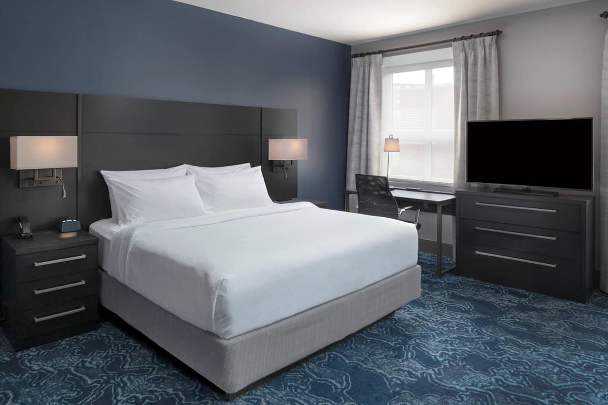 A hotel room with a large bed, bedside lamps, a desk with a chair, a flat-screen TV, and a window with curtains.