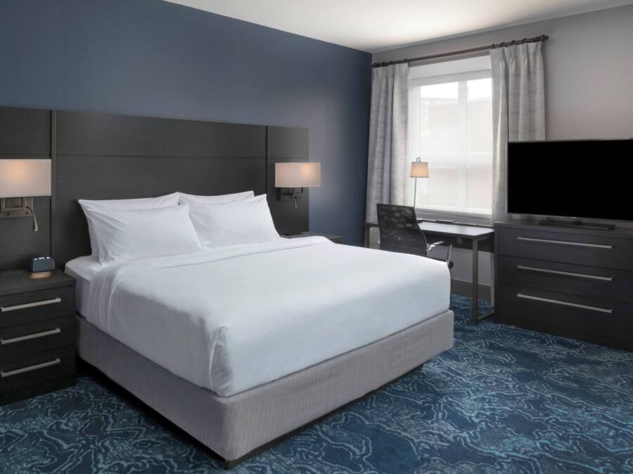 Een hotelkamer met een groot bed, nachtlampjes, een bureau met een stoel, een flatscreen-televisie en een raam met gordijnen.