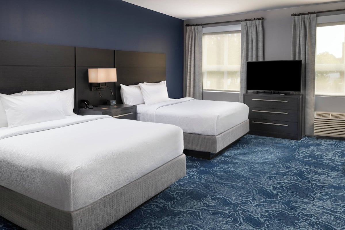 A hotel room with two beds, a flat-screen TV, a nightstand, a lamp, and a large window with curtains.