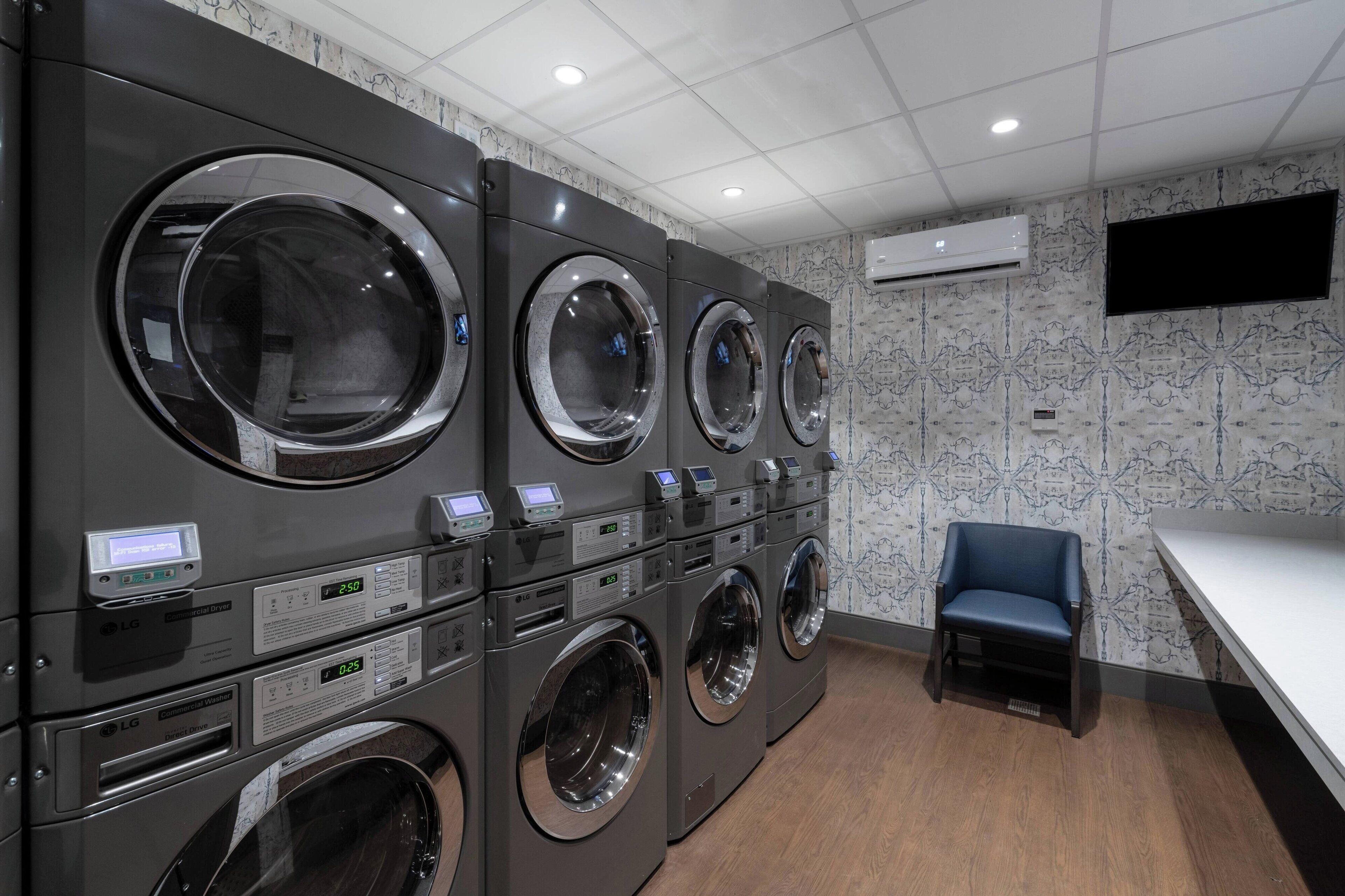 A laundry facility with multiple front-loading washing machines, a flat-screen TV mounted on the wall, and a seating area with a chair.