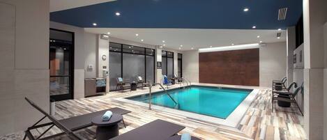 A modern indoor pool area with lounge chairs and a pool table, surrounded by a wooden floor and a blue ceiling.