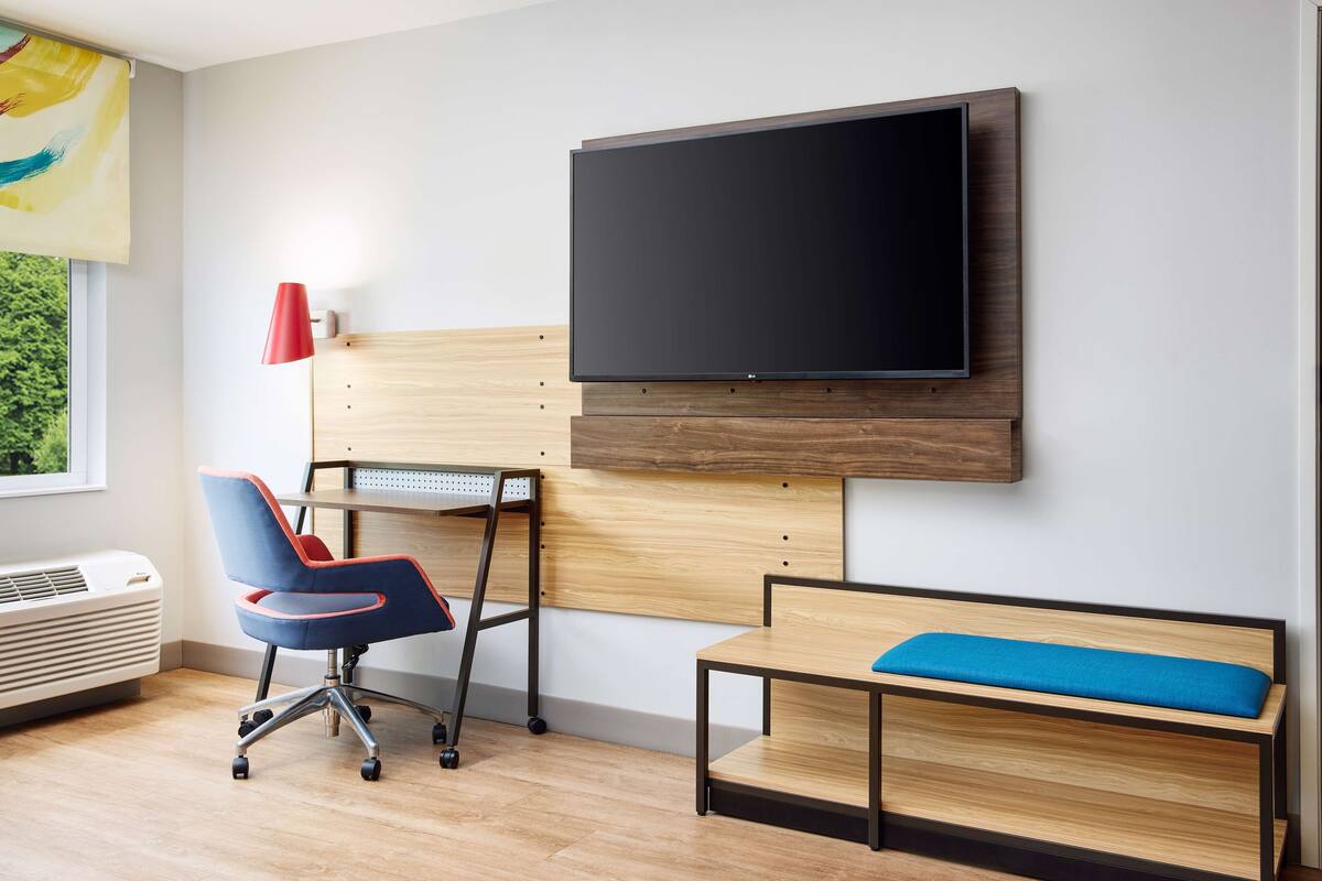 Une chambre moderne dotée d’un téléviseur à écran plat fixé au mur, d’un bureau avec une chaise, d’un banc avec un coussin et d’une lampe rouge.