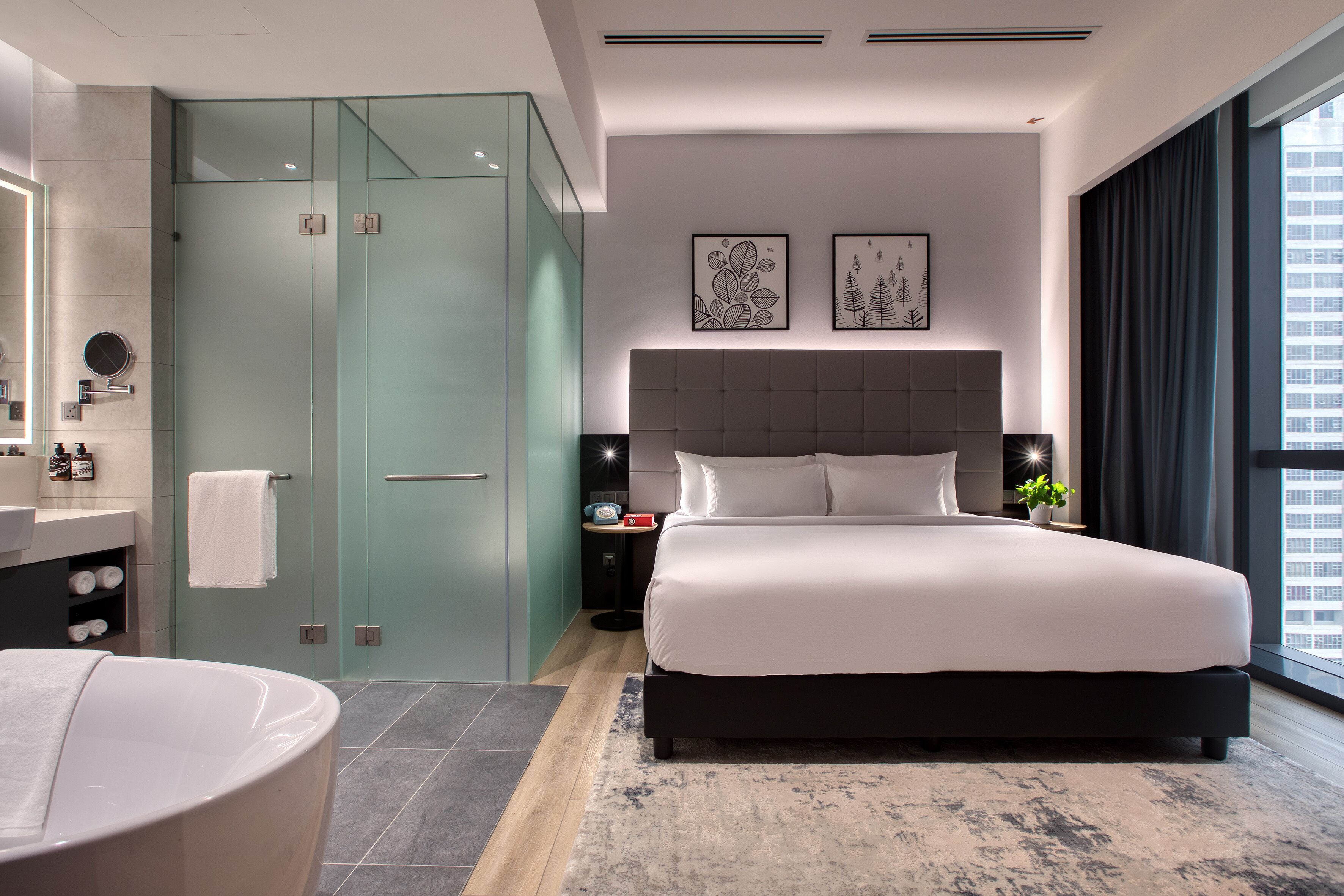 A modern hotel room with a large bed, a glass-enclosed shower, a vanity with a mirror, and a view of the city.