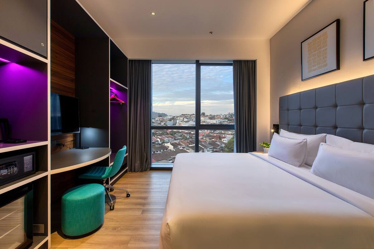 Une chambre d’hôtel moderne dotée d’un grand lit, d’un bureau avec un ordinateur, d’une chaise et offrant une vue sur la ville grâce à de grandes fenêtres.