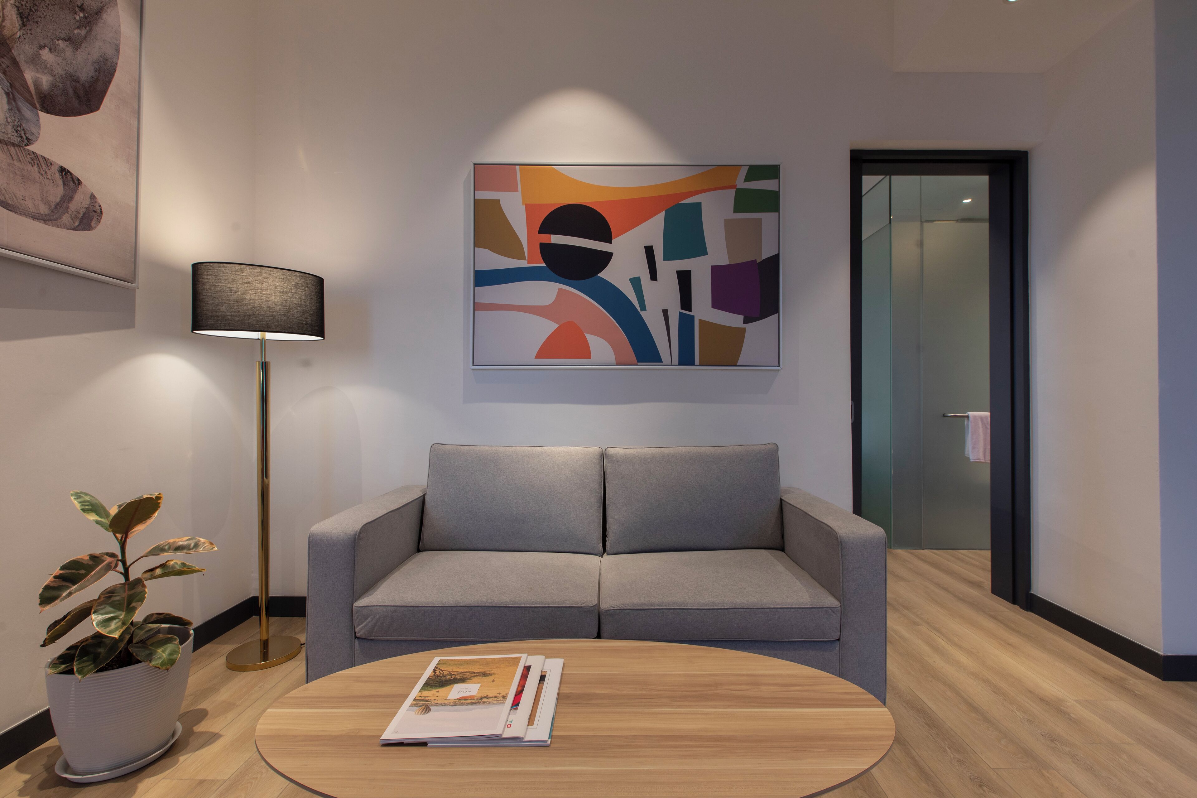 A modern interior with a grey sofa, a round wooden coffee table with a magazine, a floor lamp, and a colorful abstract painting on the wall.