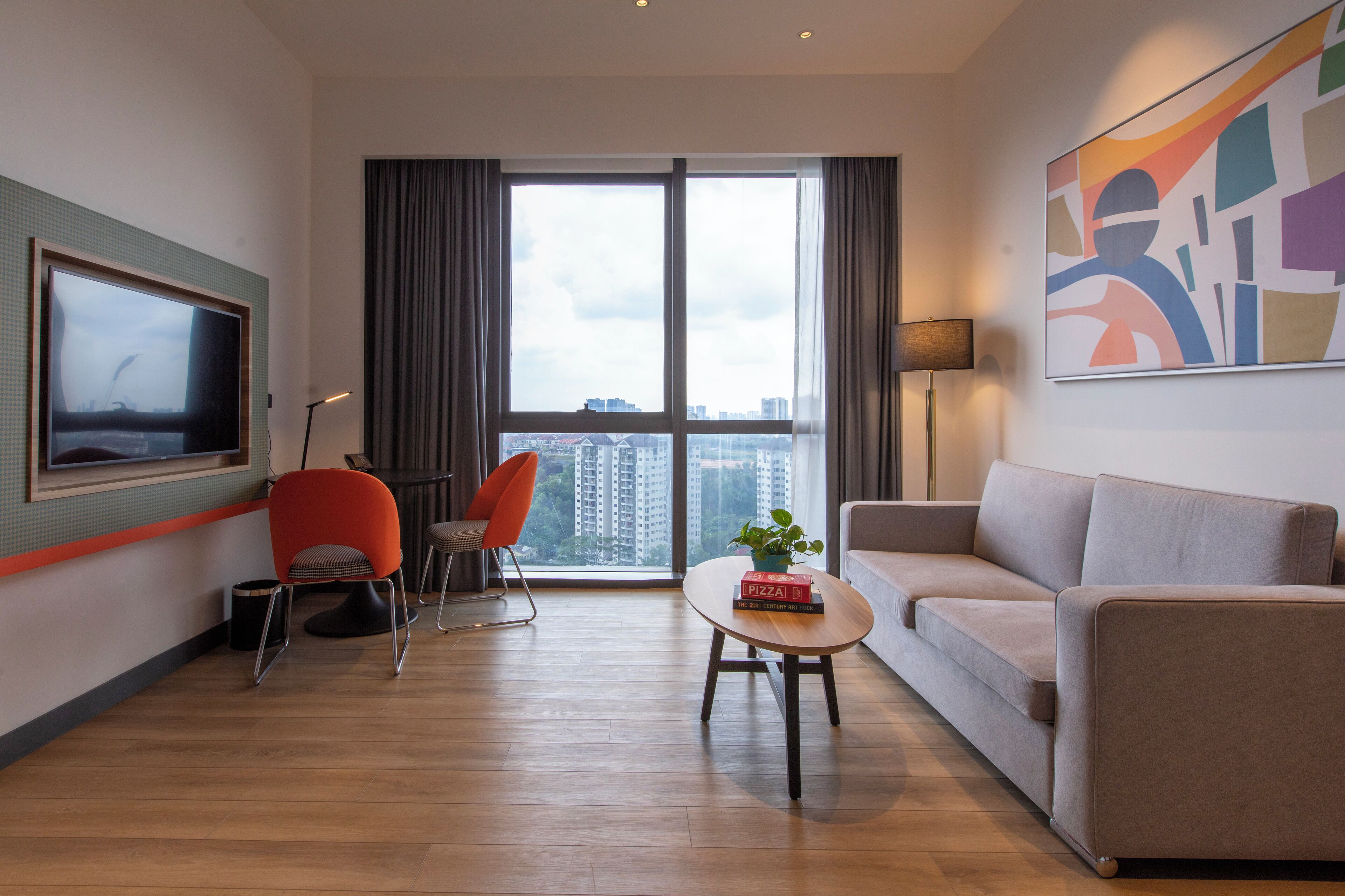 A modern hotel room with a large window, a flat-screen TV, a sofa, a coffee table, and a chair.