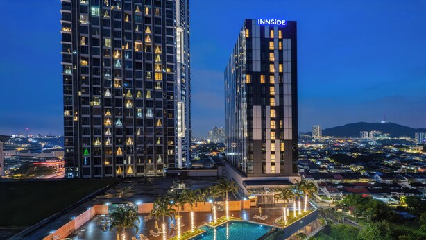 Un hotel moderno con piscina en la azotea, rodeado de exuberante vegetación y un paisaje urbano al anochecer.