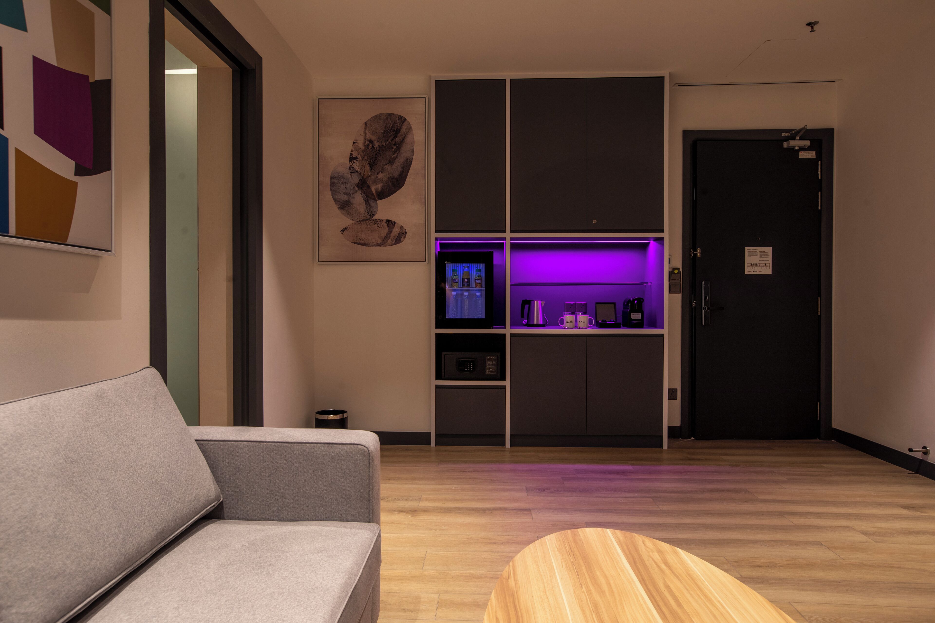 Ein modernes Wohnzimmer mit grauem Sofa, einem Holztisch und einer Küchenzeile mit Mikrowelle und Kaffeemaschine.