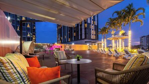 Un área de lounge en la azotea con asientos al aire libre, palmeras y vistas del horizonte de la ciudad al atardecer.