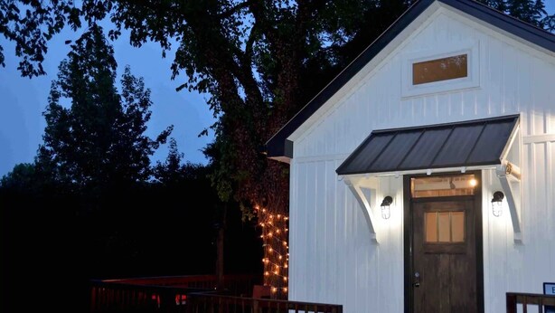 Un bâtiment blanc avec une porte en bois foncé et une petite fenêtre au-dessus. Des guirlandes lumineuses sont accrochées sur le côté du bâtiment.