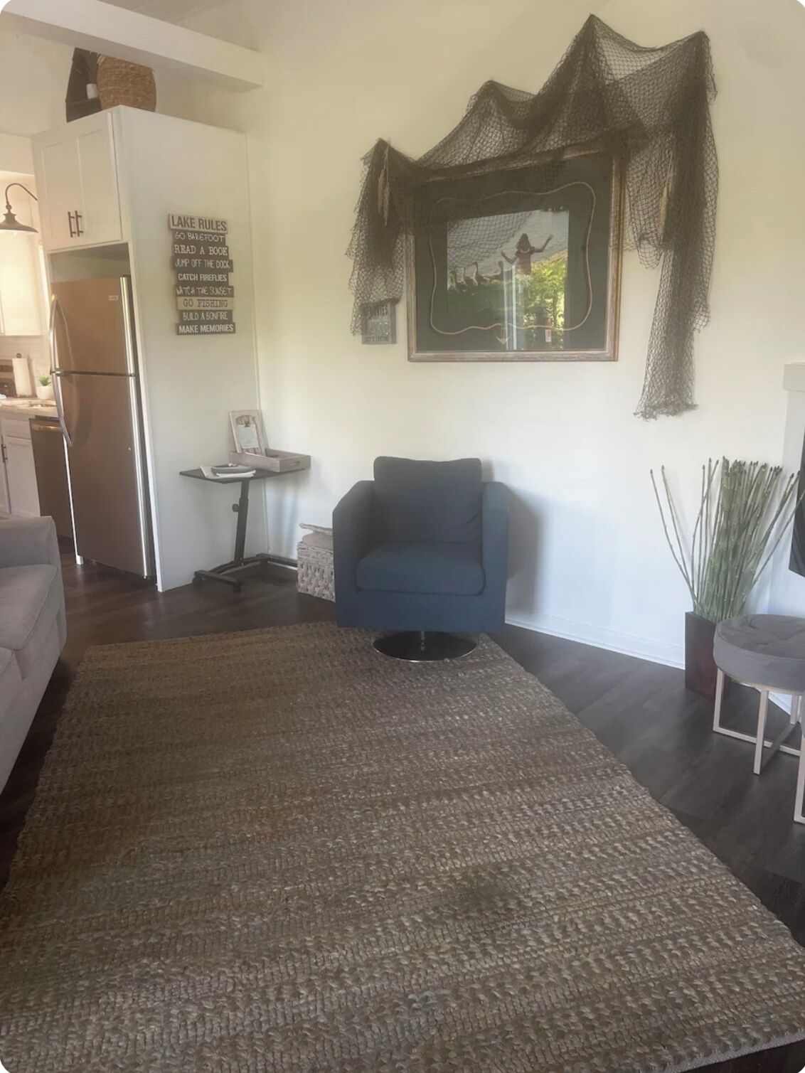 Ein Wohnzimmer mit großem Teppich, einem blauen Sessel, einem kleinen Tisch und einer Küchenzeile mit Kühlschrank.