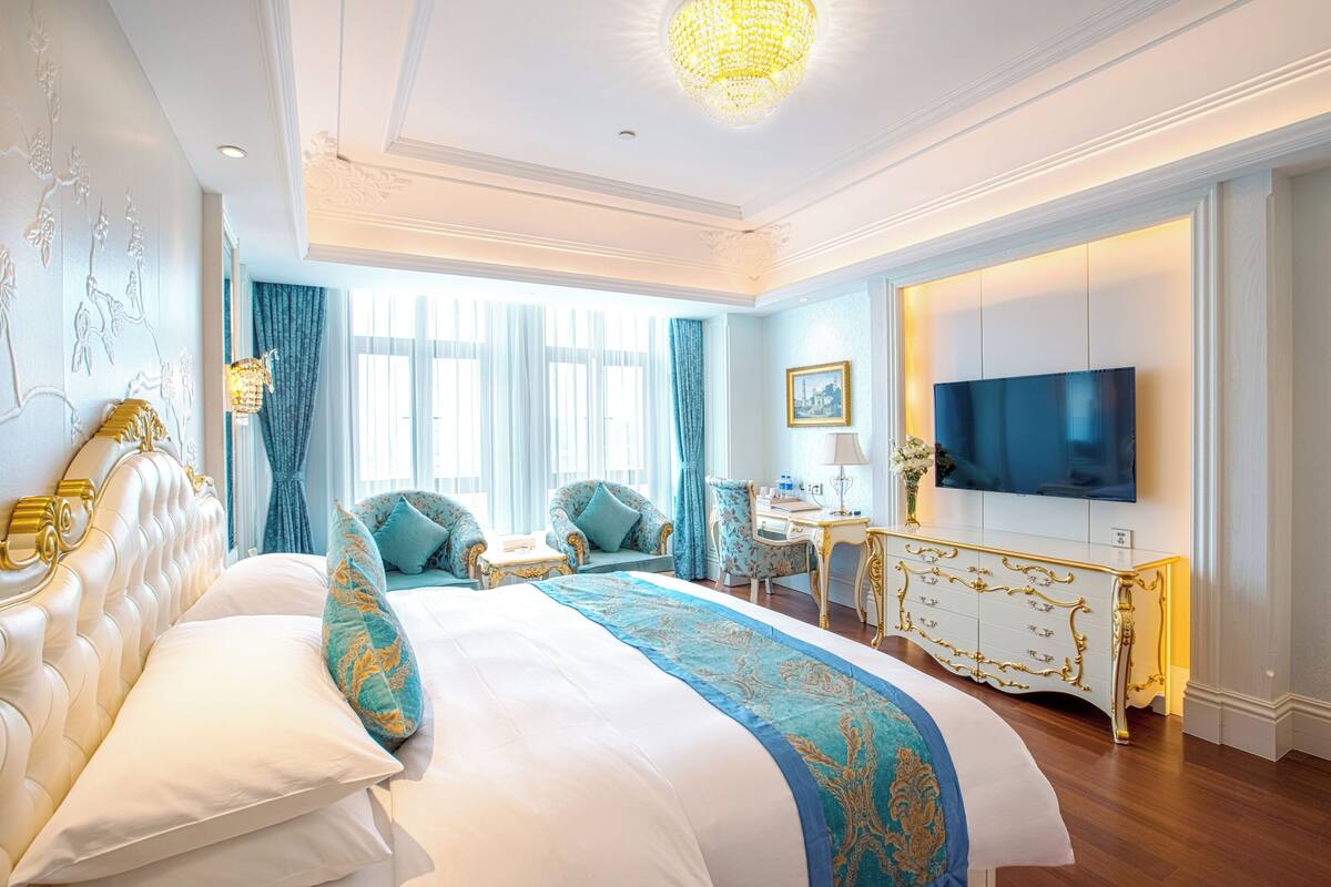 Quarto de hotel luxuoso com uma cama grande, televisão, escrivaninha e lustre.