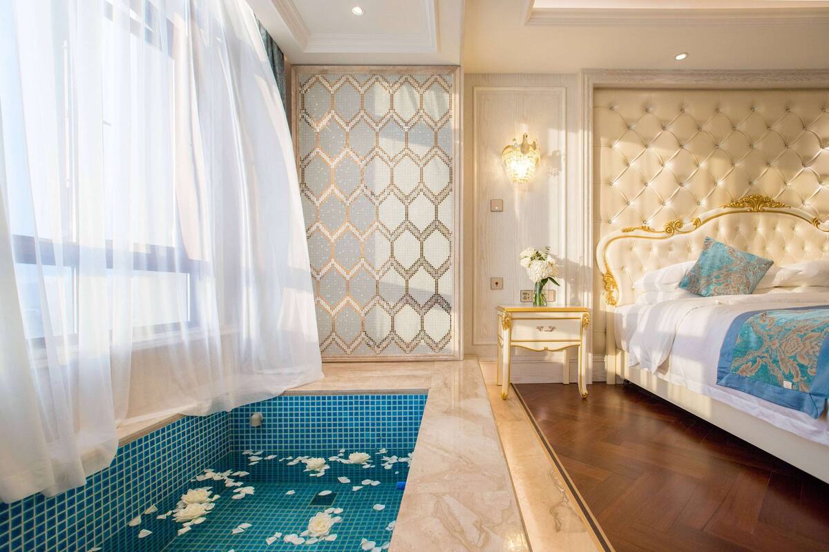 Quarto com uma cama grande, uma mesinha de cabeceira, parede decorativa e área azulejada com flores brancas.