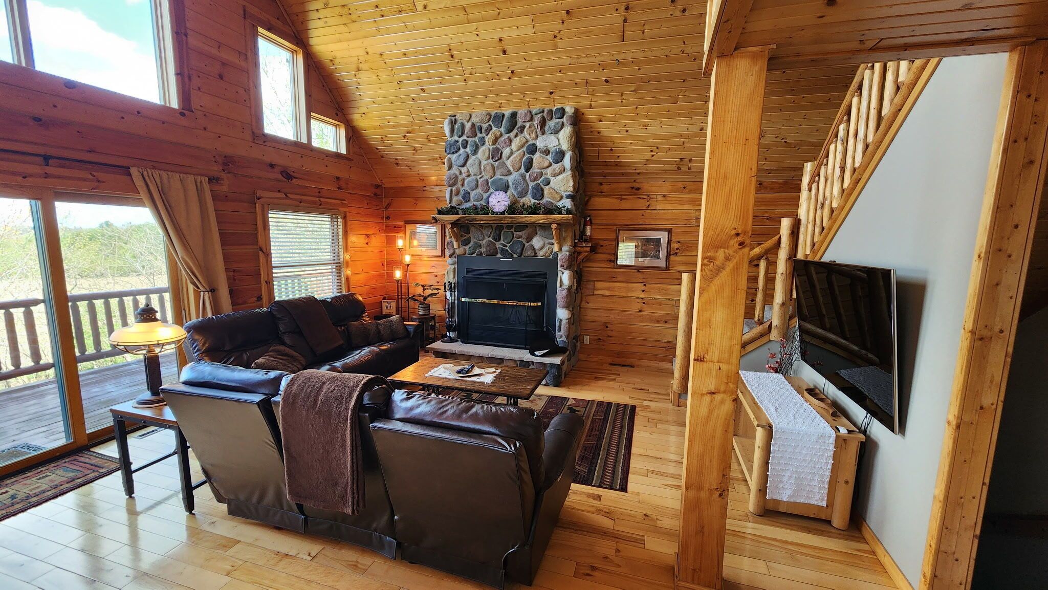 A cozy cabin interior with a stone fireplace, leather sofa, and a TV mounted on the wall.