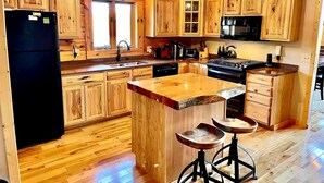 Une cuisine avec des meubles de rangement en bois, un réfrigérateur noir, une cuisinière et un évier. Il y a des tabourets près de l’îlot central et un ventilateur de plafond.