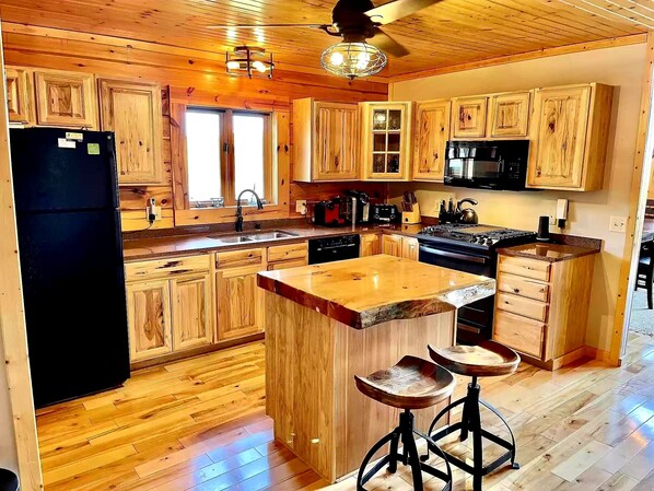 Una cocina con armarios de madera, un refrigerador negro, una estufa y un lavamanos. Hay taburetes en la isla y un ventilador de techo.