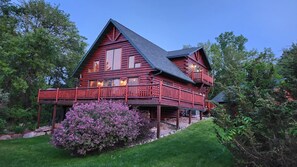 A two-story log cabin with a balcony, surrounded by lush greenery and a flowering shrub. - Devil’s Lake 5BR Cabin w/ Fireplaces,King beds,Skiing* Near Water parks in Dells (Baraboo)