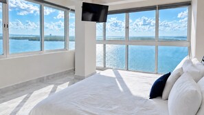 A hotel room with a large bed, a flat-screen TV, and a view of the sea through large windows.