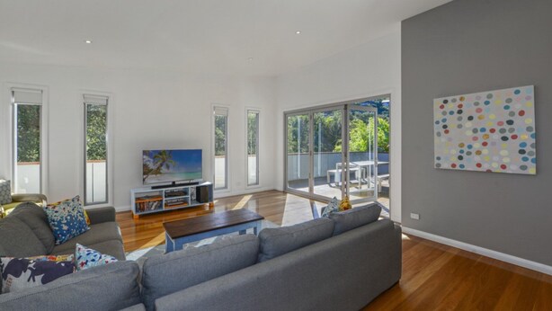 A spacious living room with a large grey sofa, a coffee table, and a TV on a stand. There are large windows and a balcony with outdoor furniture. - At The Beach - Great Family House (North Avoca)