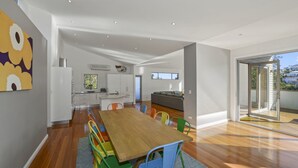 A modern kitchen with white cabinets, a dining area with a wooden table and colorful chairs, and a large sliding glass door leading to a balcony. - At The Beach - Great Family House (North Avoca)