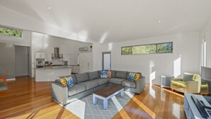 A modern living room with a grey sectional sofa, a wooden coffee table, and a view of the kitchen in the background. - At The Beach - Great Family House (North Avoca)