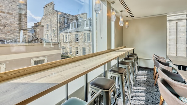 A modern interior with a long wooden counter, bar stools, and large windows offering a view of historic buildings. - Premier Inn Edinburgh Cc Waverley (Edinburgh)